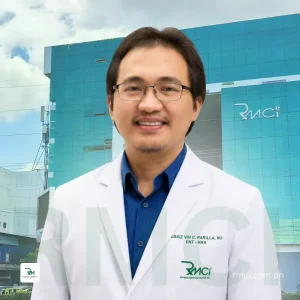 Smiling male doctor in a white coat and blue shirt standing outside a modern turquoise medical building.