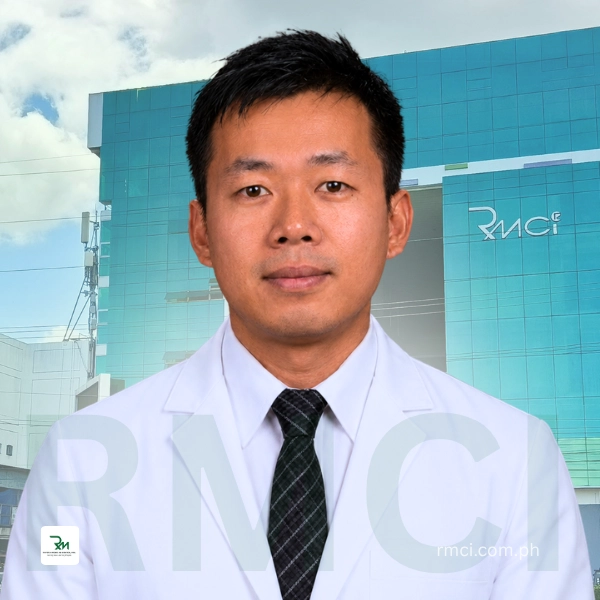 Portrait of a male doctor in a white coat and black tie standing outdoors in front of a blue RMCI building with the RMCI logo visible