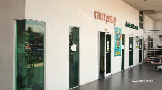 Exterior walkway of a medical clinic with an emergency entrance, glass doors, and posters along the wall on a tiled hallway