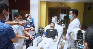 Medical team surrounding a male patient lying in a hospital bed with an IV drip, in a hallway near a CT scan room. The staff wear masks and discuss the patient’s care.