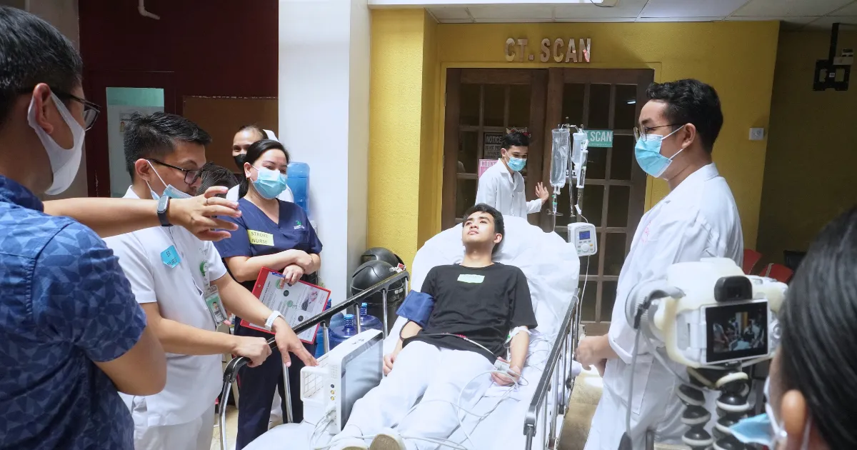 Medical team surrounding a male patient lying in a hospital bed with an IV drip, in a hallway near a CT scan room. The staff wear masks and discuss the patient’s care.